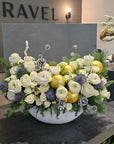 Floral arrangement with white and yellow flowers in a bowl on a dark surface, with 'RAVEL' branding in the background.