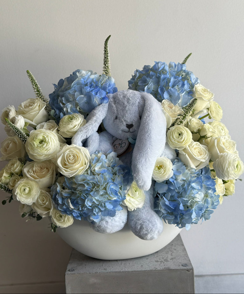 Floral arrangement with blue and white flowers and a plush toy in a white vase on a light gray background
