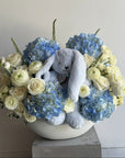 Floral arrangement with blue and white flowers and a plush toy in a white vase on a light gray background