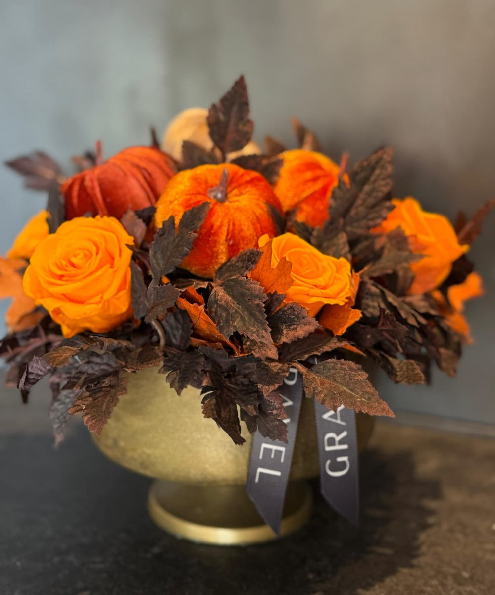 Decorative arrangement with orange flowers and pumpkins in a vase on a dark surface.