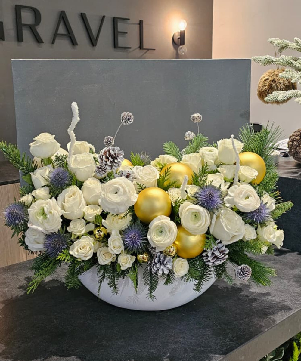 Floral arrangement with white and yellow flowers in a bowl on a dark surface, with 'RAVEL' branding in the background.